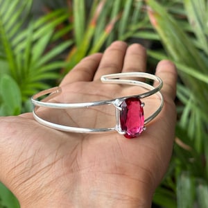 May include: A silver-toned cuff bracelet featuring a double-band design and a single, large, oval-shaped, faceted, vibrant pink gemstone set in a prong setting at the centre. The metal band is smooth and polished, creating an open-ended structure.