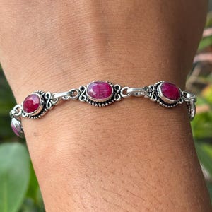 May include: A silver chain bracelet featuring multiple oval-shaped, faceted deep pink ruby gemstones. Each stone is set in a decorative silver bezel with intricate metalwork detailing, connected by small silver links to form a continuous band.