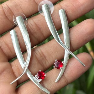 May include: A pair of silver-toned earrings featuring a textured, branch-like design with two intersecting bars. Each earring is accented with a single, round, faceted red gemstone set near the bottom curve of the metal structure.