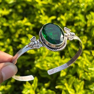 May include: A silver cuff bracelet featuring a large, oval-cut, deep green gemstone at the center. The stone is surrounded by a twisted rope-style metal border, with two small, detailed silver bee figures positioned on either side of the setting.