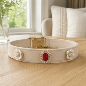 May include: A beige collar featuring a gold-toned metal buckle, a central oval-shaped red gemstone surrounded by small clear crystals, and two floral-shaped embellishments on either side, each centered with a small white pearl-like bead.