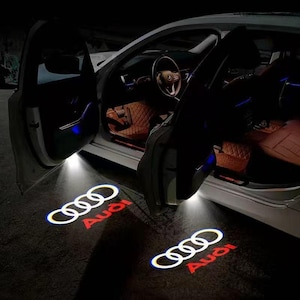 May include: A white car with open doors projecting illuminated Audi logos onto the ground. The interior features brown quilted leather seats and a steering wheel. The projected light displays the four interlocking rings and the red Audi text.