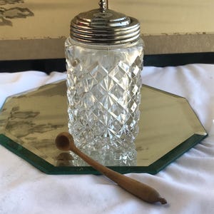 May include: A clear glass jar with a diamond pattern, topped with a silver lid. A small wooden spoon rests beside the jar. The jar is on a mirrored surface, suggesting a decorative or kitchen item.