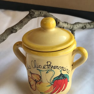 May include: A small, yellow ceramic pot with a lid and two handles. The pot is decorated with a drawing of a pitcher, garlic, and a red chili pepper. The text "Olio e Peperoncino" is written on the pot.