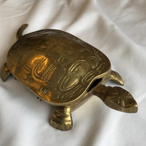 May include: A brass turtle-shaped trinket box with a hinged shell. The box is detailed with etched patterns and has a small opening. The turtle has its head and feet extended.