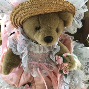 May include: A brown plush teddy bear wearing a straw hat with a wide white lace brim, a pink satin dress with white lace trim, and a small pink ribbon bow at the neckline. The bear holds a small bouquet of pink fabric roses in one paw.