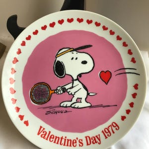 May include: A white ceramic plate featuring Snoopy playing tennis with a red heart. The plate has a pink center with red hearts around the rim and the text "Valentine's Day 1979".
