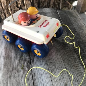 May include: A white plastic pull toy vehicle labeled Bouncing Buggy with six blue wheels featuring yellow centers. The toy includes two small figures, one with brown hair and one wearing a yellow hat, seated inside. A yellow string is attached to the front.