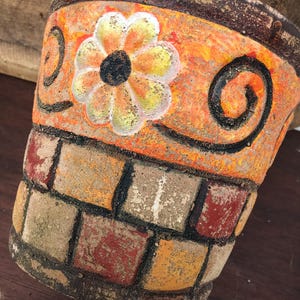 May include: Hand-painted terracotta flower pot with a vibrant orange background, a large white and yellow flower, and black swirl details. The lower section features a brick-like design in shades of red, orange, and tan. A unique decorative planter.