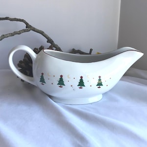 May include: White ceramic gravy boat with a handle and spout. The boat is decorated with green Christmas trees, red stars, and crescent moons. The rim is trimmed with a thin red line. A festive addition to any holiday table.