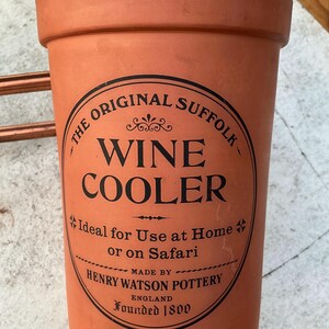May include: A terracotta wine cooler with a wide rim and a cylindrical body. The front features a black circular stamp with the text: THE ORIGINAL SUFFOLK WINE COOLER, Ideal for Use at Home or on Safari, MADE BY HENRY WATSON POTTERY ENGLAND FOUNDED 1800.