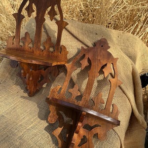 May include: Two dark brown wooden wall shelves featuring intricate cut-out patterns and a flat horizontal surface. Each shelf has a tall, decorative back panel with a pointed top and a small hole for mounting, showcasing a rich wood grain finish.
