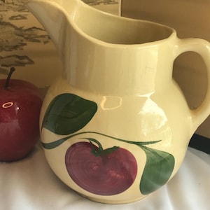 May include: Cream-colored ceramic pitcher with a rounded body and a handle. The pitcher is decorated with a large red apple design and green leaves. The pitcher has a spout for pouring and is likely used for serving beverages.
