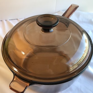 May include: A vintage amber-colored glass saucepan with a matching lid. The lid has a round knob and the pan has a handle and a small loop handle. The cookware is made of transparent glass.
