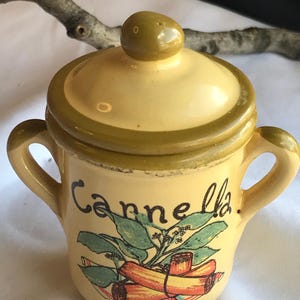 May include: A small, yellow ceramic spice jar with a lid and two handles. The jar is decorated with a graphic of cinnamon sticks and the word "Cannella". The lid and trim are olive green.