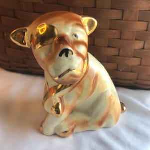 Op de afbeelding: Een keramieken beeldje van een zittende hond met een crèmekleurig lichaam en bruine markeringen. De hond heeft gouden accenten op zijn oren, poten en halsband. De snuit van de hond heeft een zwarte neus en een enigszins trieste uitdrukking. Het beeldje staat op een witte ondergrond.