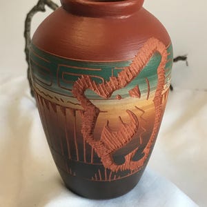 Puede incluir: Un jarrón de terracota hecho a mano con un diseño tallado. El jarrón presenta un degradado de colores, que va del marrón oscuro en la base al marrón rojizo en la parte superior. El diseño tallado es de un tono rojo más claro, con detalles en verde y amarillo.