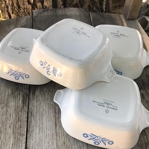 May include: Four white square ceramic pans with blue floral patterns on the sides. Each pan features two side handles and text on the bottom reading CORNING WARE P-41 PETITE PAN MADE IN U.S.A. The pans are arranged in a stacked, overlapping cluster.