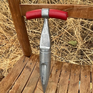 May include: A silver-colored metal garden dibber with a T-shaped handle featuring red rubber grips. The tool has a pointed tip and a textured, ribbed midsection. Embossed text on the central shaft reads "MADE IN CANADA" with a small logo above it.