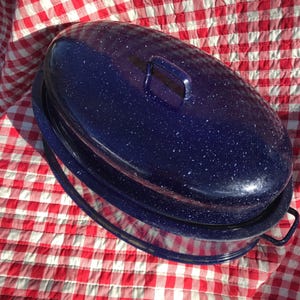 May include: A speckled navy blue enamelware roasting pan with a matching lid. The oval-shaped pan has a handle on the lid and two handles on the sides. The pan is sitting on a red and white checkered tablecloth.