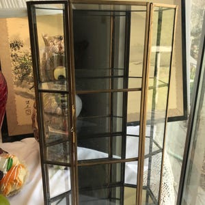 May include: A vintage glass display cabinet with a brass frame. The cabinet has multiple shelves and a hinged door. The glass panels are clear, and the frame has a warm, golden tone. The cabinet is designed to showcase collectibles.