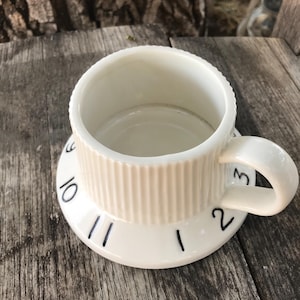 May include: A white ceramic mug with a ribbed exterior and a handle. The base of the mug features a wide, flat rim decorated with black printed numbers 1, 2, 3, 9, 10, and 11, resembling the face of a clock or a dial with markings.