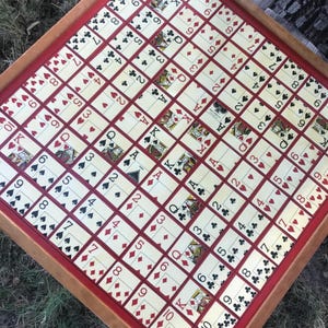 May include: A vintage card game board with a wooden frame and a red border. The playing surface is covered with a grid of playing cards, featuring various suits and numbers. The board is designed for a card game.