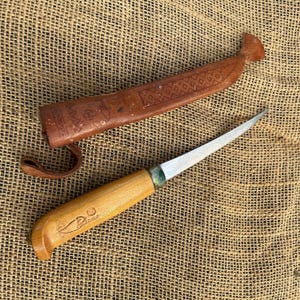 May include: A fixed-blade knife with a light-colored wooden handle featuring a stamped logo and a silver-toned metal bolster. The blade is pointed and metallic. It is accompanied by a brown leather sheath with embossed geometric patterns.