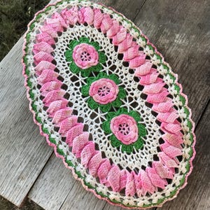 May include: An oval crochet doily featuring three central pink flowers with green leaves, surrounded by a white lacy pattern and an outer border of pink petal-like shapes. The piece is crafted with white, pink, and green yarn in a floral design.