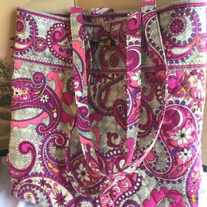 May include: A quilted tote bag with a paisley pattern in shades of pink, purple, and gray. The bag has two handles and a wooden toggle closure. The bag is made of a soft, quilted material.