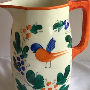 May include: Cream-colored ceramic pitcher with an orange handle and rim. The pitcher is decorated with a painted orange bird with blue wings, green leaves, and blue and orange fruit. A decorative kitchenware item.