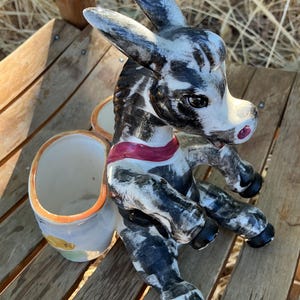May include: A ceramic donkey figurine featuring a black and white mottled coat, a red collar, and two attached cylindrical containers on its back. The donkey is in a seated position with black hooves and a small pink accent on its nose.