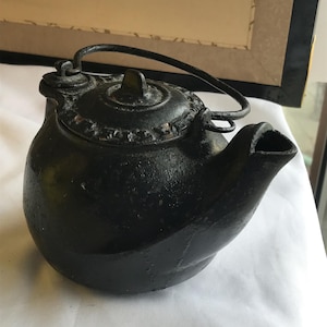 May include: A small, black cast iron teapot with a curved spout and a hinged handle. The lid is adorned with a decorative pattern. The teapot is sitting on a white surface.