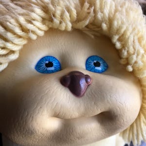 Puede incluir: Primer plano de la cara de una muñeca vintage. La muñeca tiene cabello de hilo amarillo claro, ojos azul brillante y una nariz pequeña de color marrón oscuro. La piel de la muñeca es de color amarillo pálido.