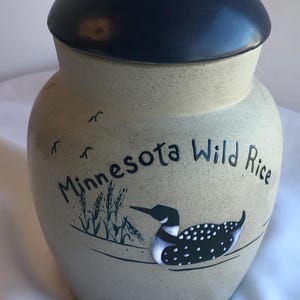 Könnte beinhalten: Ein beigefarbenes Keramikgefäß mit einem dunkelblauen Deckel. Das Gefäß ist mit den Worten "Minnesota Wild Rice", einem Eistaucher und Wildreisähren verziert. Das Gefäß wird wahrscheinlich zur Aufbewahrung von Lebensmitteln verwendet.