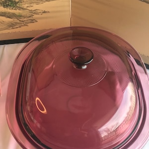 Può includere: Una pirofila rotonda in vetro color rubino con coperchio abbinato. Il coperchio ha una piccola maniglia rotonda e un design strutturato. La pirofila è in vetro trasparente.