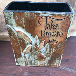 May include: A rectangular ceramic vase with a black interior and a textured exterior. The vase features a brown and tan color scheme with a botanical design and the words "Take time to live".