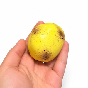 May include: A small, rounded, bright yellow object with a smooth surface texture, featuring several irregular dark brown to black speckled patches distributed across its exterior, resembling a stylized or artificial fruit.
