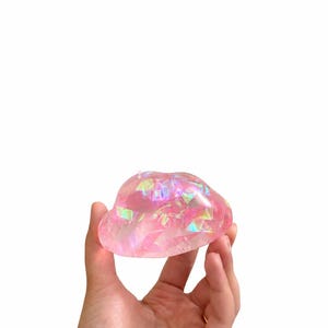 May include: A translucent, rounded object with a pink hue, featuring embedded iridescent, holographic shards that reflect light in various colors. The surface is smooth and polished, creating a glossy appearance with a soft, organic, cloud-like shape.