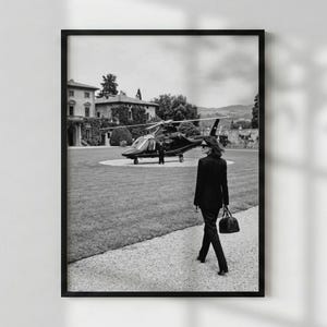 May include: A black and white photograph featuring a person in a dark blazer and trousers walking toward a helicopter parked on a grassy lawn. The scene includes a large stone building in the background, with the person carrying a dark handbag.