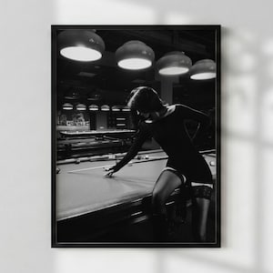May include: A black and white photograph of a person in a long-sleeved black dress and lace-trimmed stockings leaning over a pool table. The scene is set in a dimly lit room with several large pendant lights hanging above the billiard tables.