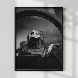 Op de afbeelding: Een zwart-wit ingelijste print met een piloot die een helm met een sterembleem draagt, in een cockpit zit en een handleiding leest met de tekst HOW TO FLY op de omslag. De scène is zichtbaar door de gebogen glazen cockpitkap.