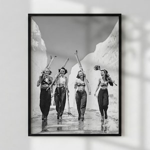 May include: A black and white framed photograph depicting four individuals walking through a narrow path between high snowbanks. Each individual is wearing a bikini top and dark trousers, carrying skis and poles while smiling in a snowy landscape.