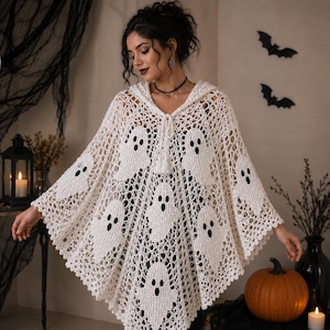May include: A cream-colored crochet hooded poncho featuring a repeating pattern of white ghost motifs with black embroidered eyes and mouths. The garment has an open-weave design, a drawstring neckline with tassels, and a scalloped hemline edge.