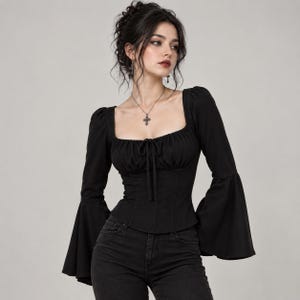 Può includere: Un top nero a maniche lunghe con scollo quadrato e laccetti centrali, corpetto aderente e maniche a campana svasate. Il capo è abbinato a jeans in denim scuro e a una collana con ciondolo a croce in argento su una catenina sottile.