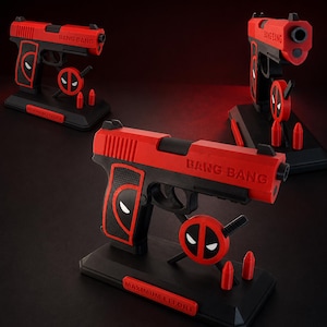 May include: A red and black toy pistol display stand featuring the text BANG BANG on the barrel and MAXIMUM EFFORT on the base. The stand includes a circular logo with white eyes and two small red bullet-shaped accessories on a black base.