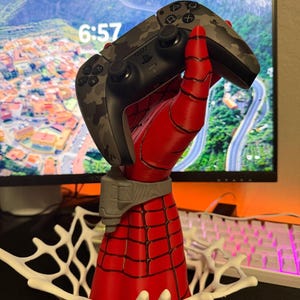 May include: A red and black hand sculpture with a web-like pattern holding a gray camouflage gaming controller. The sculpture features a gray wrist cuff and is supported by a white, web-shaped base, creating a display stand for the controller.