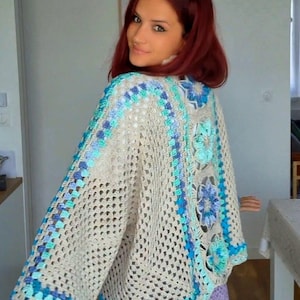 Peut inclure: Un gilet en crochet beige avec un motif à mailles ouvertes et des manches larges. Le vêtement est agrémenté d'un panneau vertical de motifs floraux en relief dans des tons bleu clair, bleu foncé et turquoise, bordé de fil bleu assorti.