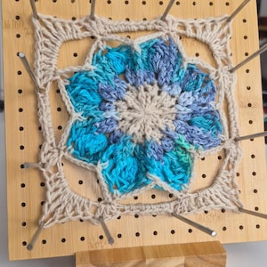 Peut inclure: Une planche de blocage carrée en bois avec une grille de trous et des tiges métalliques maintenant un carré au crochet. La pièce présente une étoile centrale crème entourée de pétales dans des tons de bleu, turquoise et violet, bordée de fil crème.