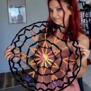 Peut inclure: Une décoration murale circulaire en crochet présentant un motif de toile noir à l'extérieur avec des accents intérieurs en fil orange, jaune, rose et violet. Le design rayonne depuis un point central, créant une structure géométrique ajourée.